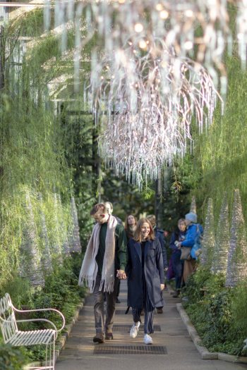 Two people stroll down a long hallway with plants along the walls and white chandeliers hanging from the ceiling