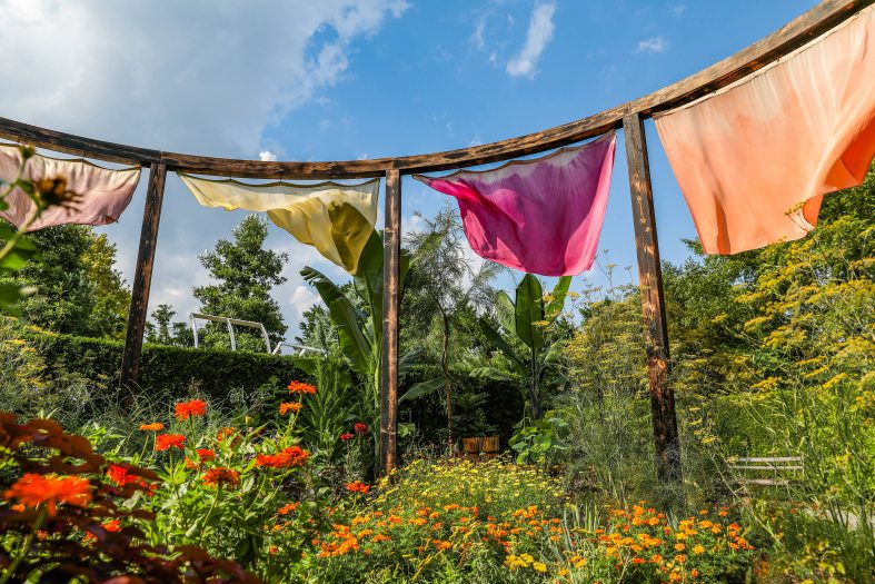 A tall wooden structure with hanging pieces of fabric ripple in the wind over a bed of green plants and small orange flowers