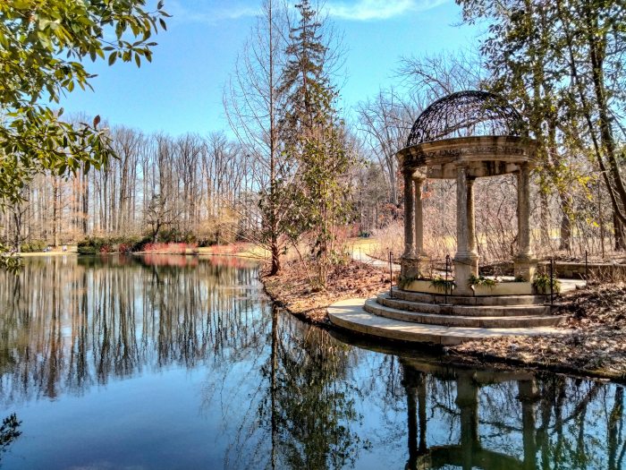 A stone decorative structure stands on the edge of a large lake with dormant winter trees surrounding