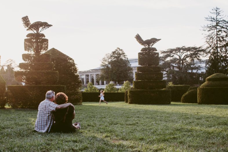 A couple sits on the ground surrounded by shaped topiary with a child running in the distance during sunset