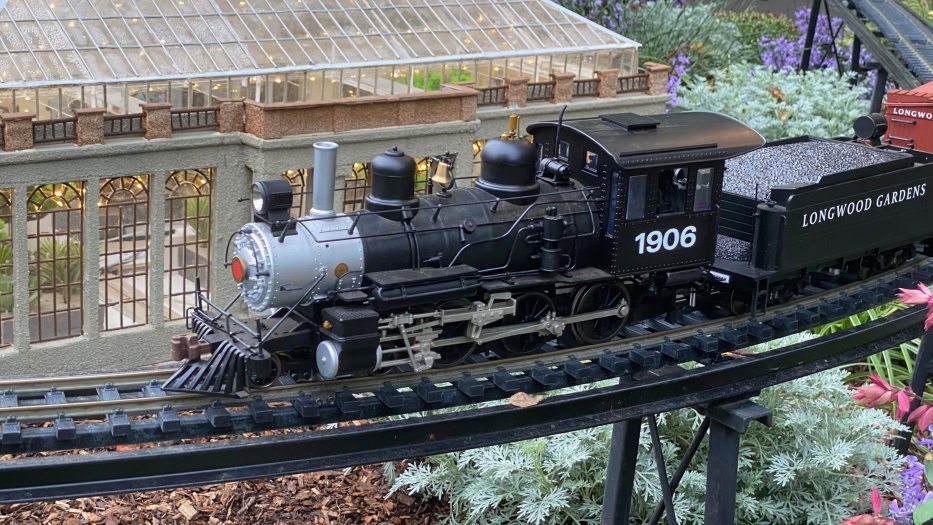 A black and silver miniature locomotive labeled 1906 pulls a coal car labeled Longwood Gardens past a model Conservatory through an outdoor garden.