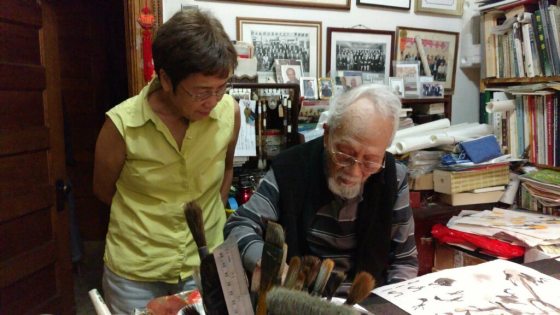 two people at a desk with one watching the other paint