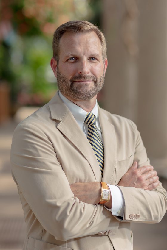 Headshot of a person in a beige suit and arms crossed looking at the camera.