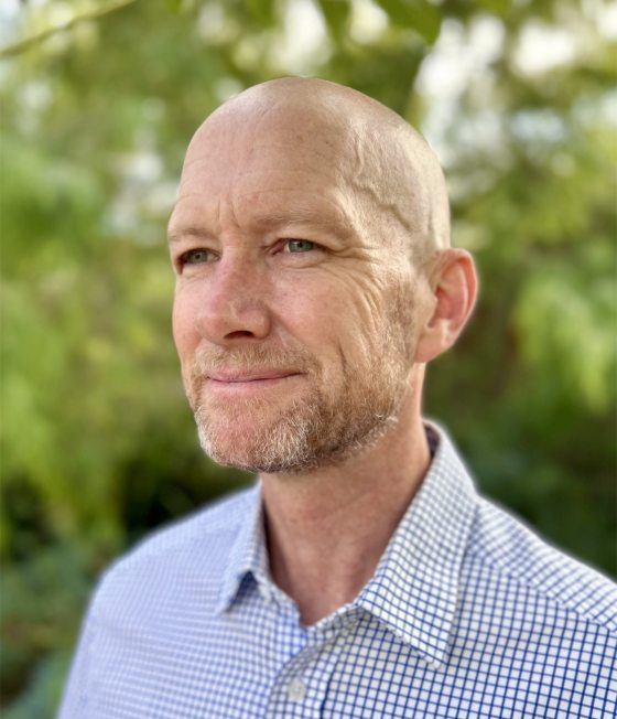 A headshot of a white man in a checkered blue pattern shirt with trees in the background 