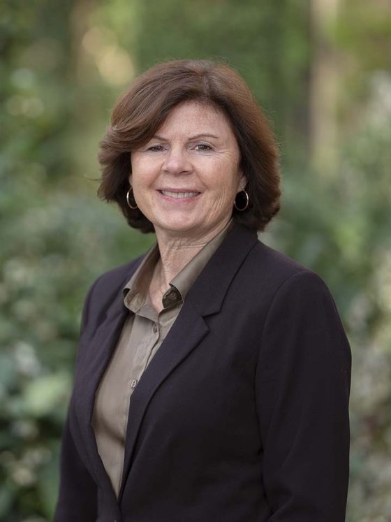 A person with reddish-brown collar-length hair and medium hoop earrings smiles at the camera, wearing an olive green collared shirt under a dark suit jacket buttoned at the waist.