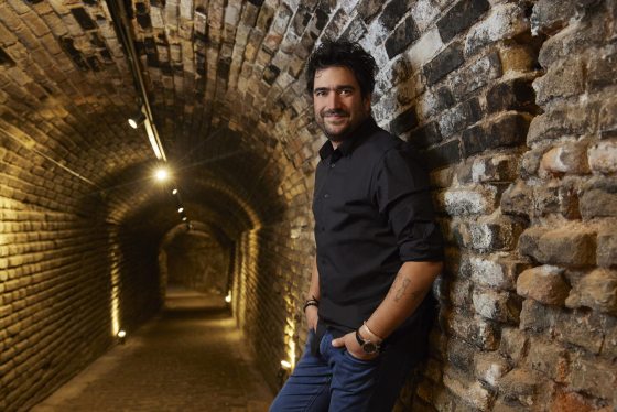 A person in a black shirt and jeans, leaning against a stone wall and smiling at the camera.