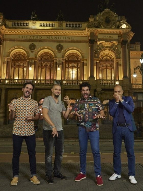 Four musicians mime playing invisible instruments in front of an ornate brightly lit building at night.