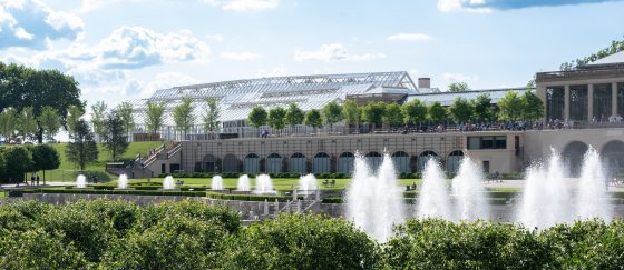 Fountains can be seen in the forefront of the photo. In the background, a new glass conservatory can be seen on top of an arched lower façade and next to another building.  