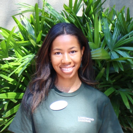 Portrait of smiling person with medium-toned skin and long black hair parted down the middle, wearing a dark green Longwood Gardens t-shirt.