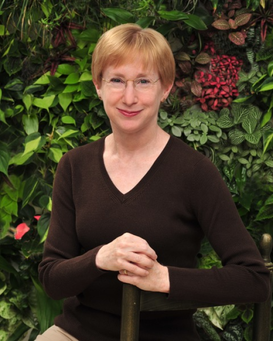 A smiling person with short reddish-blond hair, wearing glasses and a dark brown V-neck sweater, poses with hands folded on the back of a chair.