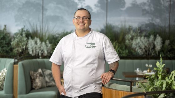 A person in a white chef coat resting against a sage green couch in the middle of the 1906 restaurant.