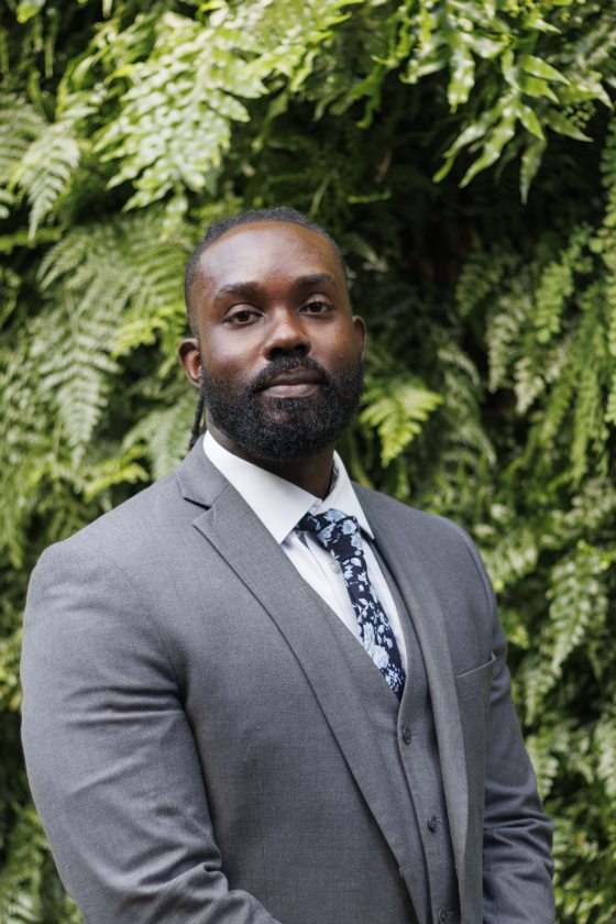 A black man in a grey suit with a white shirt and blue tie poses in front of a leaf-covered wall. 