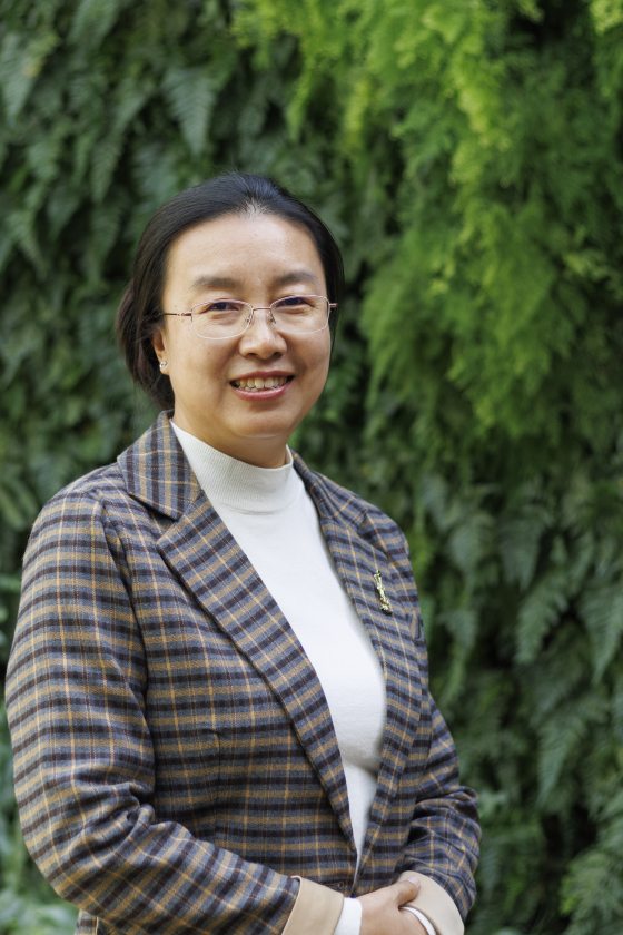 A woman with black hair and glasses wearing a plaid grey jacket stands in front of a leaf-covered wall. 